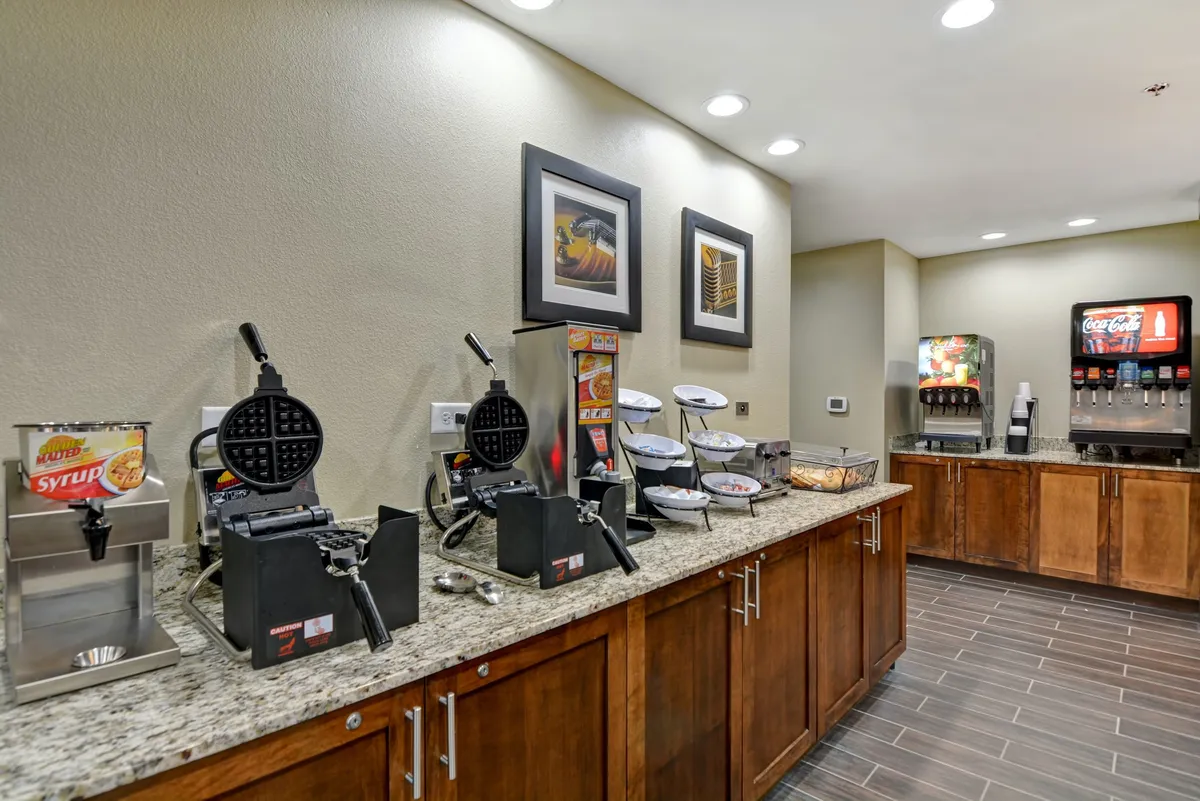 This image features a granite countertop with a waffle maker, syrup dispenser, and a beverage station set against a backdrop of framed artwork, all well-lit by morning light. The setup exudes a casual, inviting atmosphere, ideal for enjoying a hotel breakfast buffet. The elements present highlight the variety and self-service nature of the complimentary breakfast offering.
