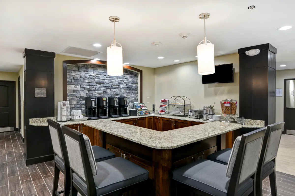 A bright kitchenette area with a granite countertop, modern coffee machines, a stone wall accent, stylish hanging lights, and a visible seating area. The space is organized for both preparing light meals and enjoying coffee, creating a welcoming and communal environment.
