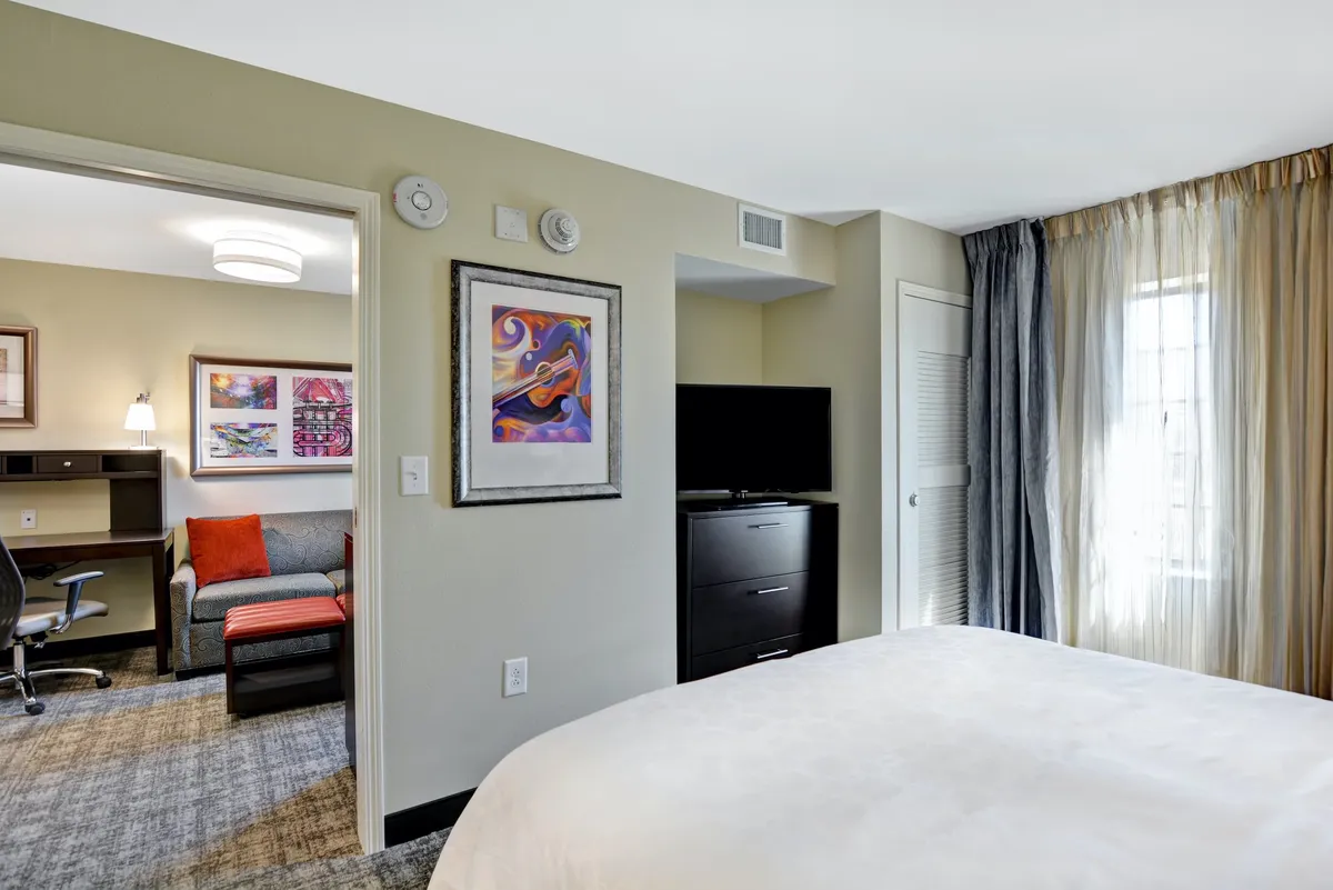 This image showcases a comfortable living area featuring a bed, an inviting couch, wall-mounted television, and tasteful artwork. Natural light streams in through the window, and the space is styled for both relaxation and productivity.