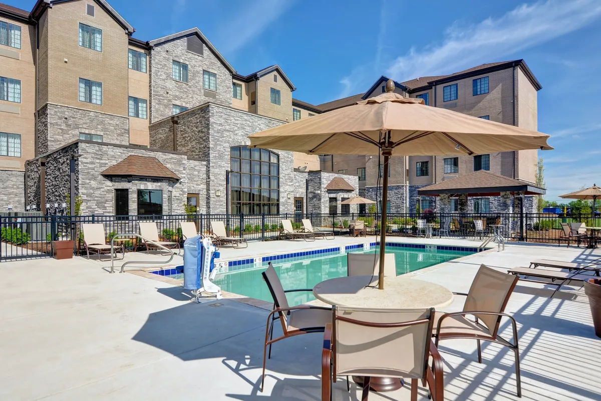 This image captures the hotel's inviting outdoor swimming pool, surrounded by lounge chairs and umbrellas. The building's exterior provides a welcoming backdrop, while guests can be seen swimming, sunbathing, and enjoying meals together poolside. The relaxed, social setting is perfect for families unwinding after a day of exploring local parks and attractions.