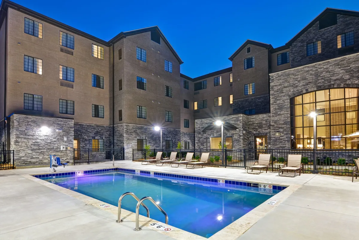 A vibrant outdoor scene with a sparkling swimming pool lined with lounge chairs and set against the building facade. Soft outdoor lighting creates a welcoming, relaxed atmosphere, perfect for swimming or enjoying sunset gatherings.