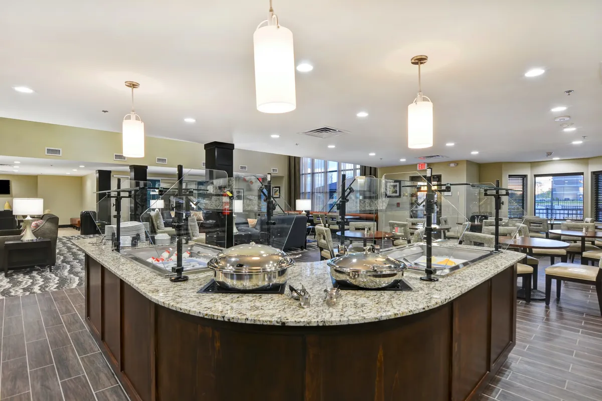 A bright, communal food service area is depicted, featuring a well-organized seating arrangement and expansive windows that fill the space with natural light. The overall mood is communal and inviting.