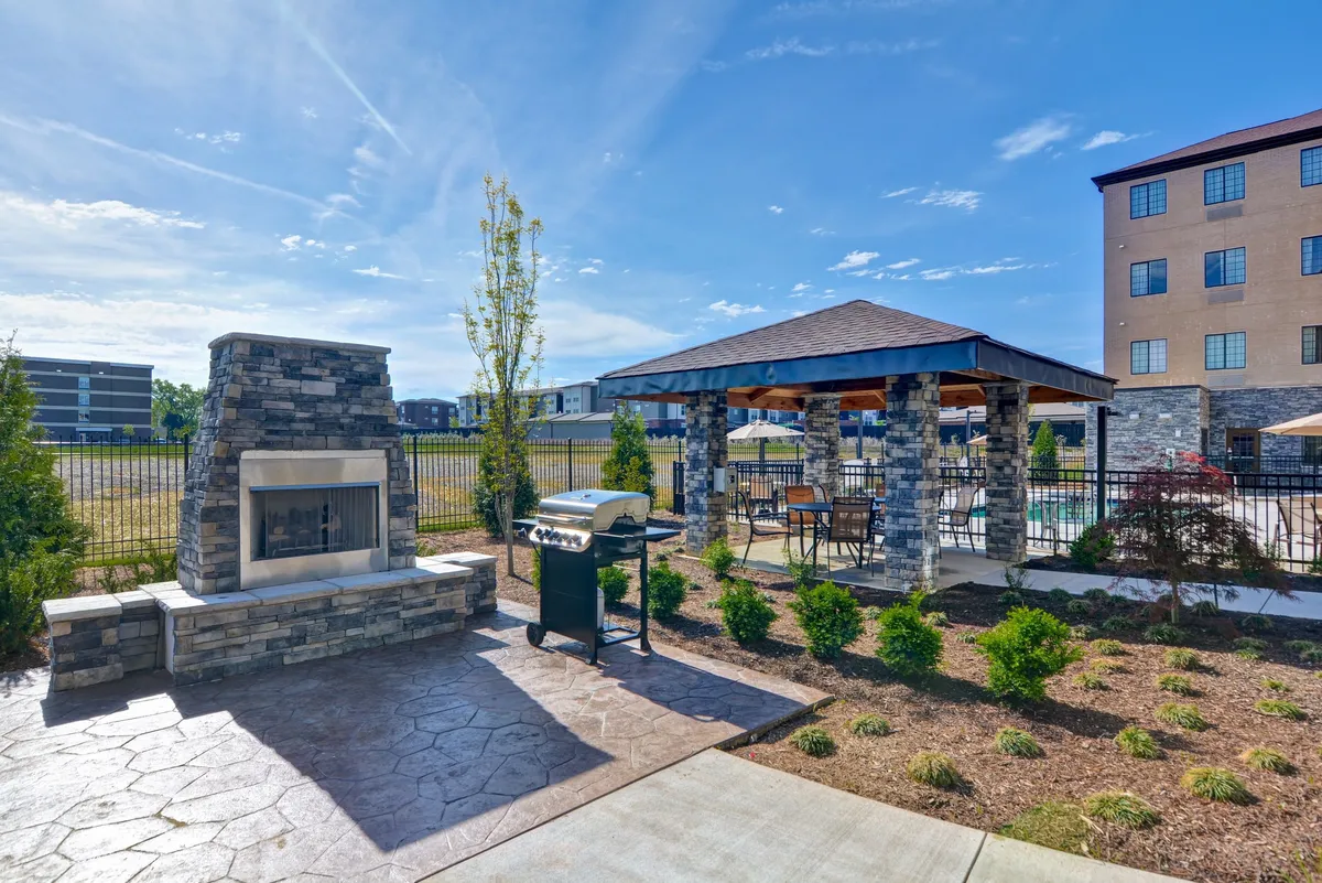 The exterior of the property featuring a welcoming stone fireplace, grill, gazebo, and lush landscaped garden. People gather around the fire, cook outdoors, and enjoy a relaxed, social setting, evoking a sense of community and opportunity for informal networking.
