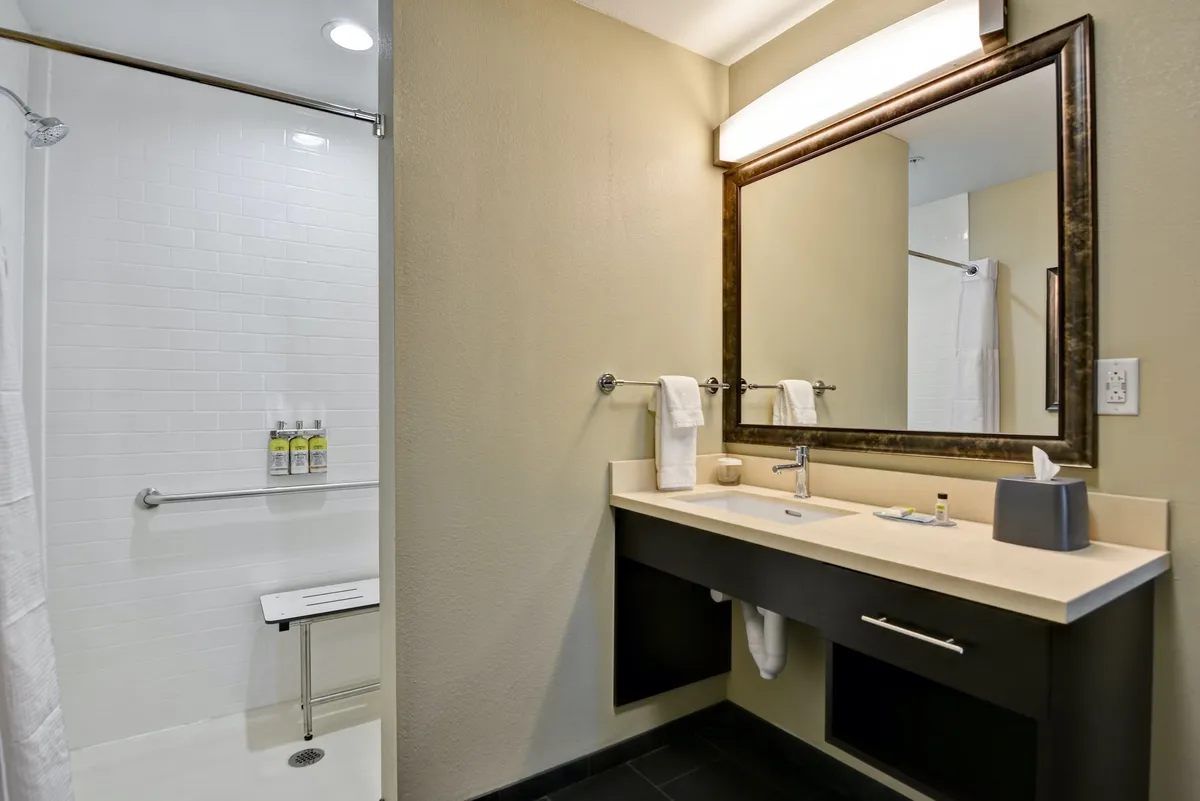 A bright and serene bathroom interior featuring a modern sink with a large mirror, fresh white towels neatly arranged, and a glass-enclosed shower. The space exudes cleanliness and is set up for personal grooming and relaxation after a long day.