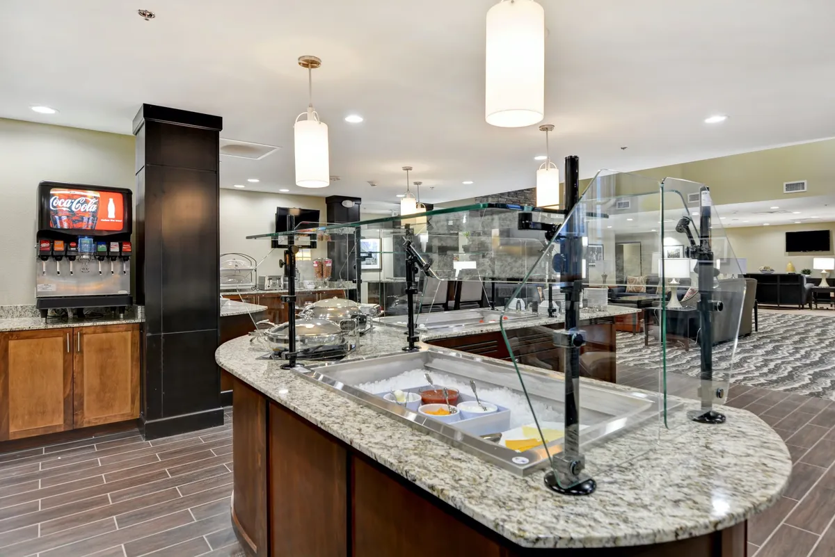 A food display area with a granite countertop, beverage station, and hanging light fixtures, designed for self-service dining. The mood is relaxed and friendly, with a layout that encourages socializing over shared meals in a spacious, well-lit setting.