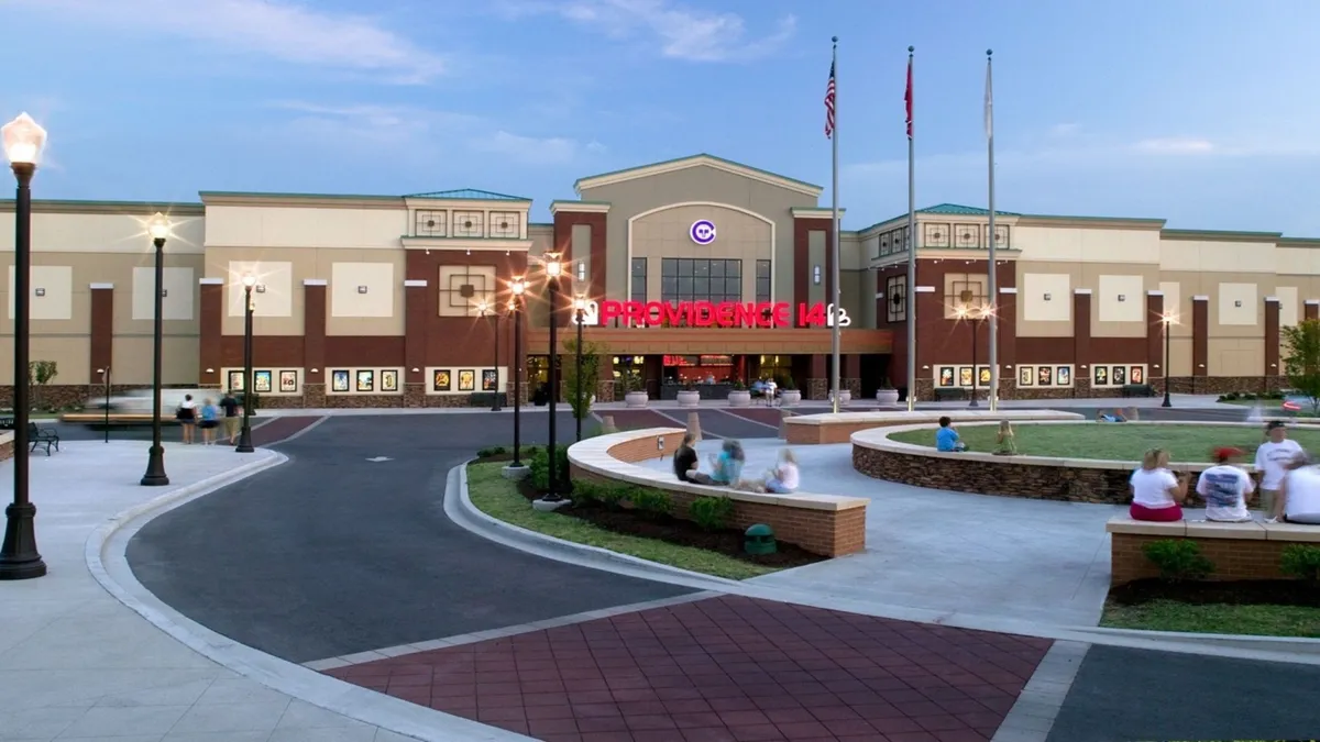 The image shows the lively entrance of a local theater, highlighted by a bright, illuminated marquee and surrounded by a seating area around a fountain. People are socializing and enjoying the communal atmosphere, suggesting both entertainment and networking opportunities nearby.
